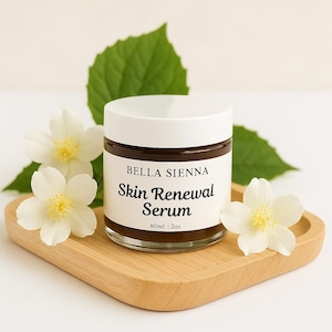 May include: A glass jar of Bella Sienna Skin Renewal Serum with a white lid, set on a wooden tray. The label reads "Skin Renewal Serum" and "60ml | 2oz". White flowers and green leaves surround the jar.