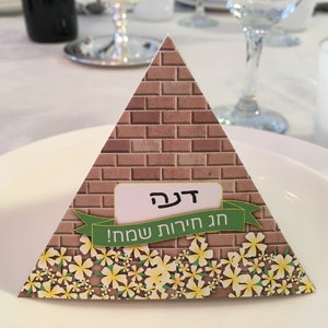 May include: A triangular place card with a brick pattern and a green ribbon. The ribbon has the text "Happy New Year!" in Hebrew. The place card has a white name tag with the name "Dena" written in Hebrew.