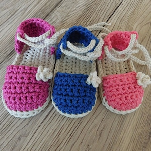 May include: Three handmade crochet baby sandals in pink, blue, and pink. The sandals have a white sole, straps, and a small flower accent. The sandals are displayed on a light wood surface.
