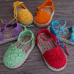May include: Six handmade crochet sandals in various colours. The sandals have a neutral sole with bright uppers in yellow, orange, purple, green, red, and blue. Each sandal has a strap for securing the foot.