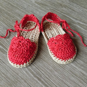 Op de afbeelding: Een paar handgemaakte gehaakte sandalen. De sandalen hebben een rood, geschulpte ontwerp op de teen en enkelbandjes, met een natuurlijke beige zool. De sandalen worden vastgebonden met rode koorden.