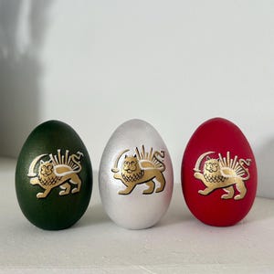 May include: Three decorative Easter eggs in green, silver, and red. Each egg features a gold-coloured lion and sun emblem. The eggs are displayed on a white surface, with a potted plant in the background.
