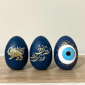 May include: Three blue eggs decorated with gold paint. The first egg features the Iranian lion and sun symbol. The second egg has gold calligraphy in Farsi. The third egg has a blue and white evil eye design.