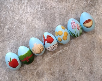 May include: Seven blue painted eggs with different designs. The designs include a fish, a garlic clove, a green patch of grass, an apple, a bowl of rice, a flower, and a pile of coins.