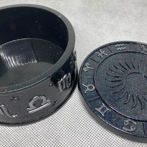 Puede incluir: Caja de baratijas de resina negra con tapa a juego. La caja y la tapa están decoradas con símbolos del zodiaco plateados y un diseño de sol. La caja es redonda y tiene un acabado brillante.