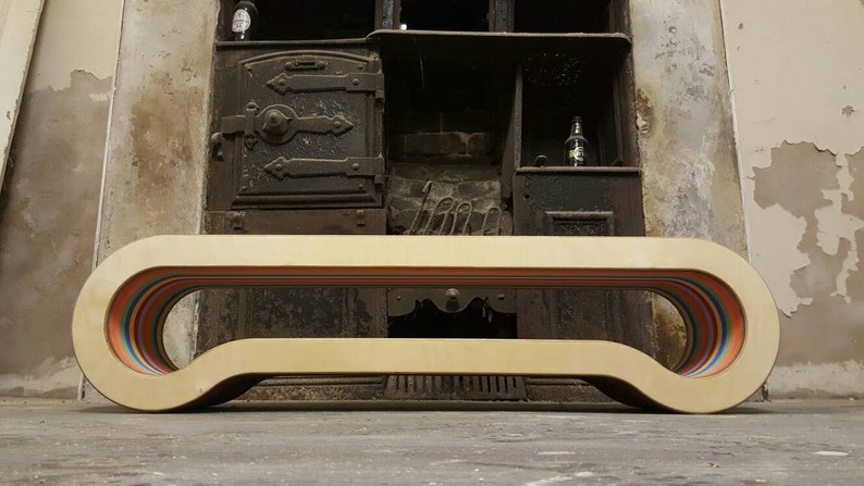 May include: A light wood bench with a curved design and a rainbow stripe detail. The bench is in front of a vintage fireplace.