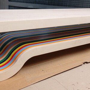 May include: A white wooden bench with a unique design featuring a curved, multi-colored, striped underside. The bench has a modern, minimalist aesthetic.