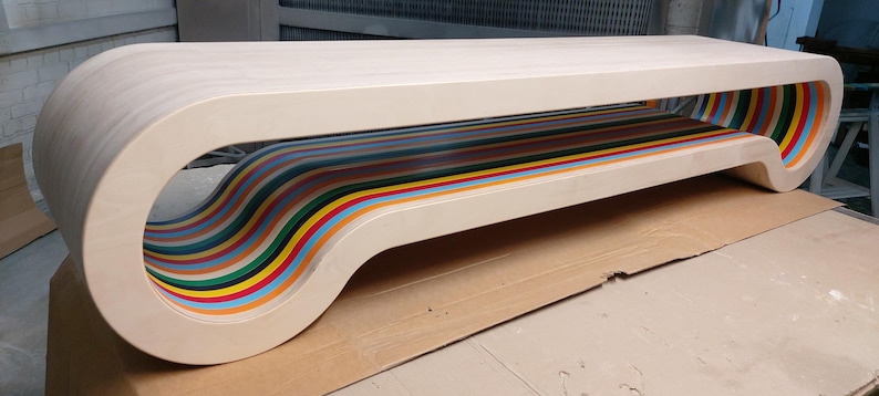 May include: A white wooden bench with a unique design featuring a curved, multi-colored, striped underside. The bench has a modern, minimalist aesthetic.