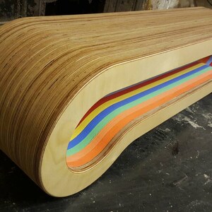 May include: A wooden bench with a rainbow-colored cutout design. The bench is made of plywood and has a curved shape.