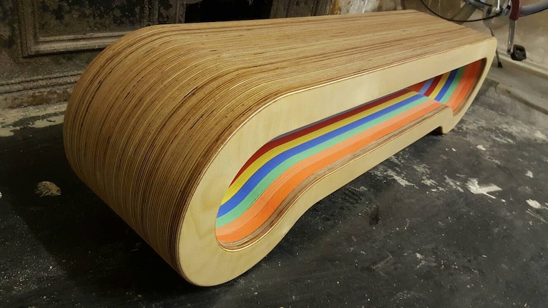 May include: A wooden bench with a rainbow-colored cutout design. The bench is made of plywood and has a curved shape.