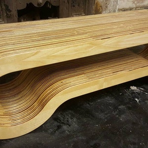 May include: A modern, wooden bench made of layered plywood, with a unique curved design. The bench has a smooth, light brown finish and a rounded, open shape.