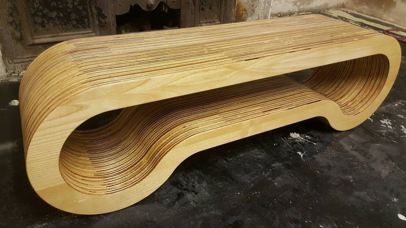 May include: A modern, wooden bench made of layered plywood, with a unique curved design. The bench has a smooth, light brown finish and a rounded, open shape.