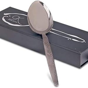 May include: A stainless steel serving spoon with a patterned handle rests on a black box. The spoon has a large, oval bowl and a textured handle. The box has white text and a stylized design. The spoon is a kitchen utensil.