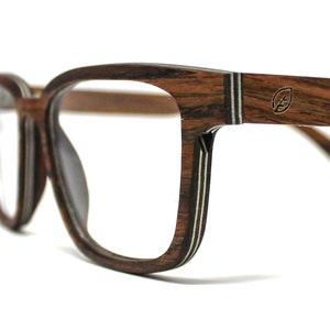 K&ouml;nnte beinhalten: Brille mit einem dunkelbraunen Holzmaserungsrahmen, der eine geschichtete Konstruktion mit sichtbaren hellen Streifen an den Kanten aufweist. Der Brillenb&uuml;gel zeigt ein kleines metallisches blattf&ouml;rmiges Logo-Emblem nahe dem Scharnier.