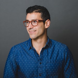May include: A person wearing a dark blue button-down shirt with a white polka dot pattern and brown-rimmed eyeglasses. The shirt features a standard collar, a front button placket, and a single chest pocket on the left side of the garment.