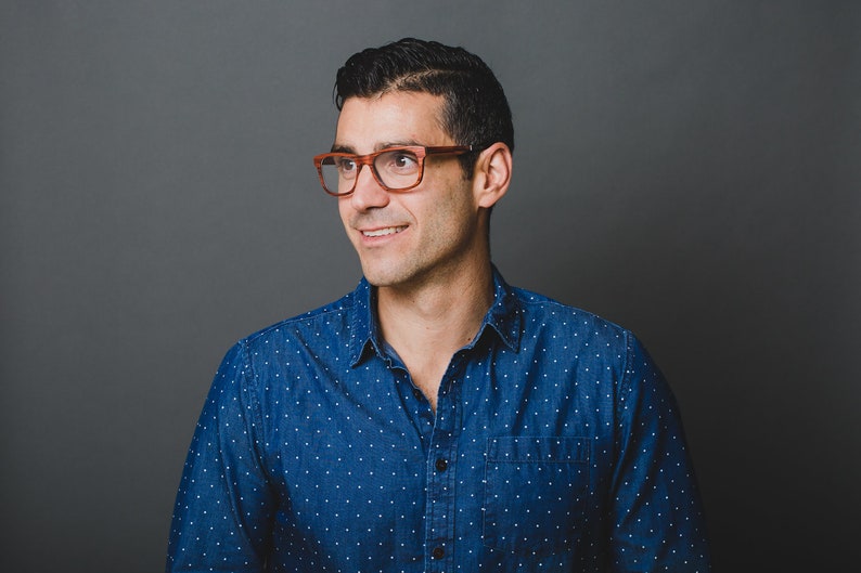 May include: A person wearing a dark blue button-down shirt with a white polka dot pattern and brown-rimmed eyeglasses. The shirt features a standard collar, a front button placket, and a single chest pocket on the left side of the garment.