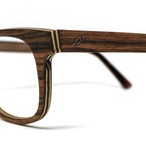 May include: Eyeglasses featuring a frame with a layered wood grain pattern in shades of brown and tan. The temples display a small circular logo with a leaf icon. The temple tips are black, and the frame includes clear lenses for vision correction.