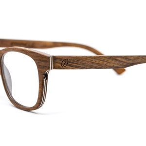 May include: Eyeglasses with a dark brown wood grain frame featuring a layered construction visible on the edges. The temples display a small circular logo with a leaf icon. The clear lenses are set within a rectangular frame shape.