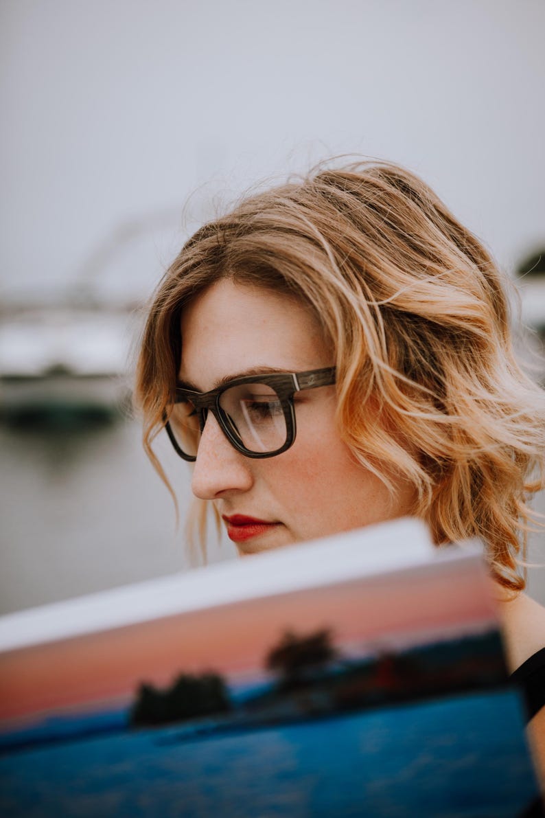May include: A pair of dark-toned, rectangular-framed glasses with a visible wood grain texture. The frames feature a straight top edge and rounded lower rims, resting on a face with blonde hair and red lipstick while holding a colorful printed image.