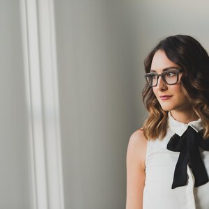 K&ouml;nnte beinhalten: Eine &auml;rmellose wei&szlig;e Bluse mit einer schwarzen Schleife am Kragen, kombiniert mit einer rechteckigen Brille mit dunklem Rahmen. Das Kleidungsst&uuml;ck hat eine durchgehende Knopfleiste und einen strukturierten Kragen, getragen von einer Person mit schulterlangem, welligem braunem Haar.