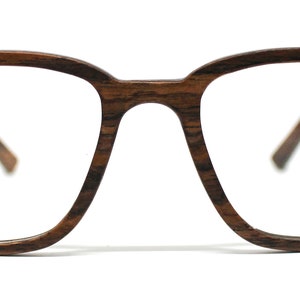 K&ouml;nnte beinhalten: Brille mit einem rechteckigen Rahmen in dunkelbrauner Holzmaserung. Der Rahmen verf&uuml;gt &uuml;ber einen Steg, der die beiden Gl&auml;ser verbindet, sowie zwei B&uuml;gel, die sich zu den Seiten erstrecken. Das Design zeigt eine nat&uuml;rliche Holzstruktur.