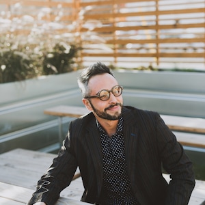 May include: A dark pinstriped blazer worn over a navy blue button-down shirt with a white polka dot pattern. The attire is paired with round-framed glasses featuring a brown tortoiseshell finish and dark blue denim jeans.