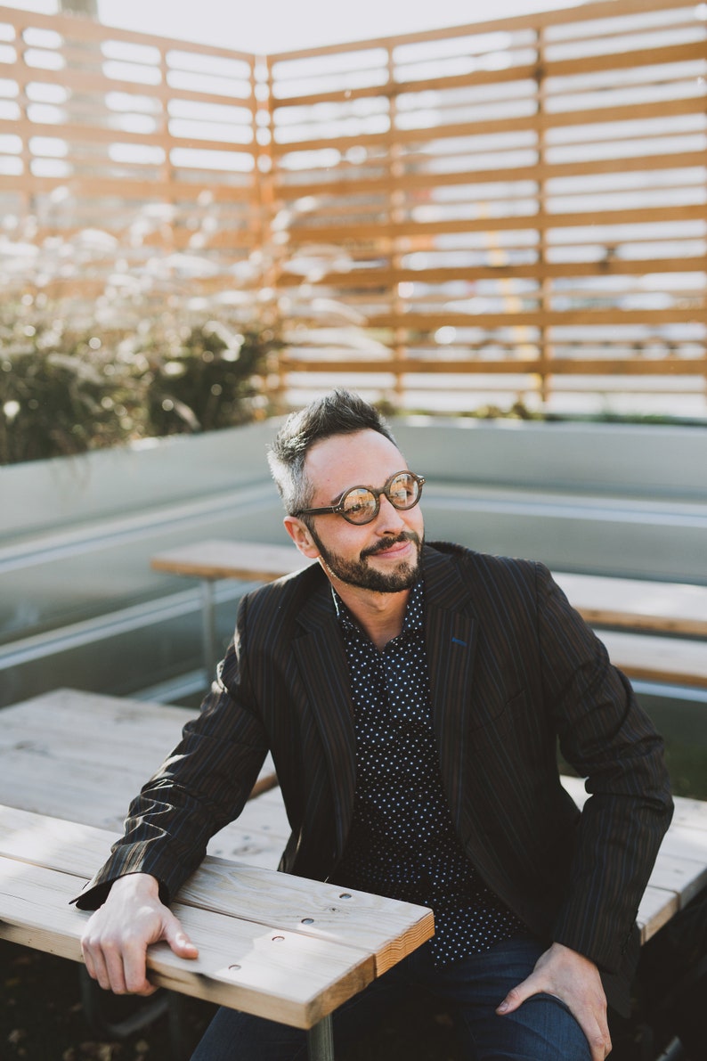 May include: A dark pinstriped blazer worn over a navy blue button-down shirt with a white polka dot pattern. The attire is paired with round-framed glasses featuring a brown tortoiseshell finish and dark blue denim jeans.