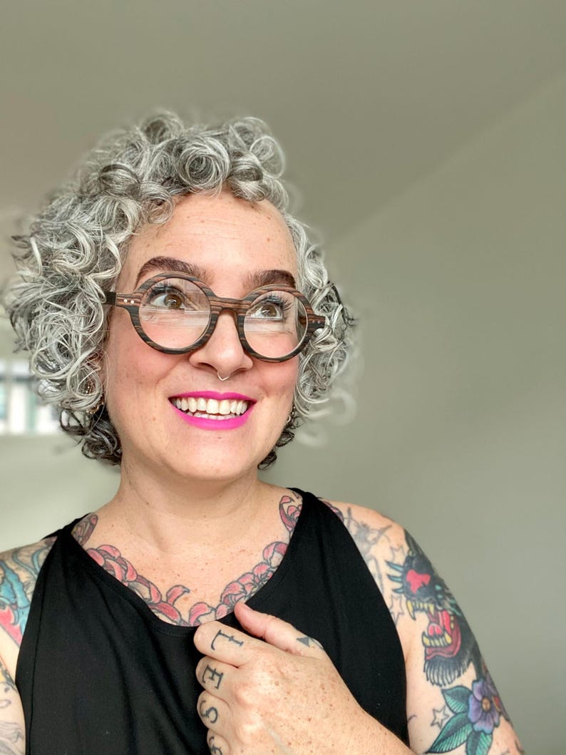 May include: Round eyeglasses featuring a dark brown and black wood-grain textured frame. The glasses have a circular shape with a visible horizontal striped pattern on the rims and small silver-toned metal accents on the outer corners of the frame.