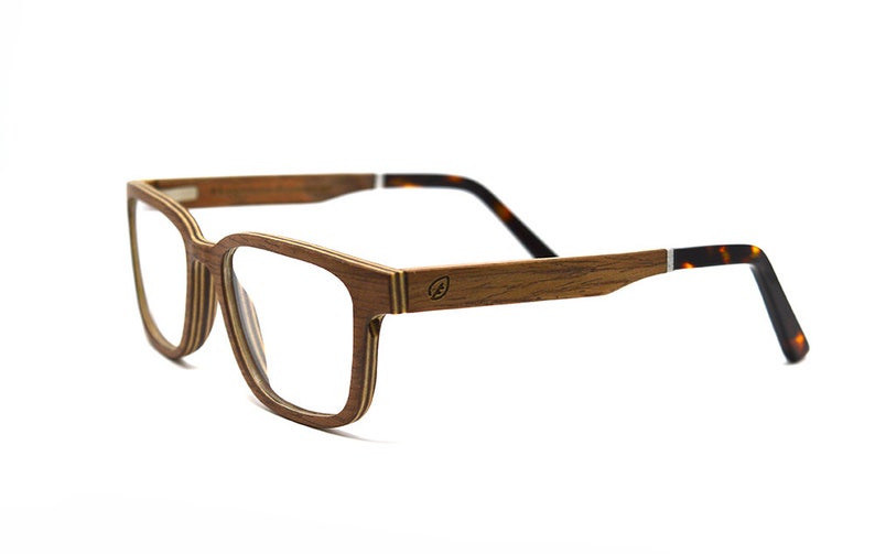 K&ouml;nnte beinhalten: Brille mit einem geschichteten Holzrahmen mit sichtbarer Maserung. Die B&uuml;gel bestehen aus Holz und gehen an den Enden in ein Schildpattmuster &uuml;ber. Ein kleines kreisf&ouml;rmiges Logo ist an der Au&szlig;enseite des B&uuml;gels eingraviert.