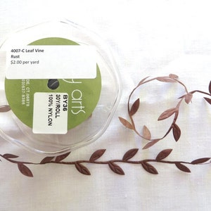 May include: A spool of rust-colored leaf vine with the text "4007-C Leaf Vine Rust" and "30Y/ROLL 100% NYLON". The vine is made of nylon and is displayed on a white surface. The leaf vine is a decorative craft supply.