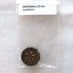 May include: A gold and black button, 22 mm in diameter, with an ornate floral design. The button is in a clear plastic bag with a label that reads "2010 Button, 22 mm Gold/Black".