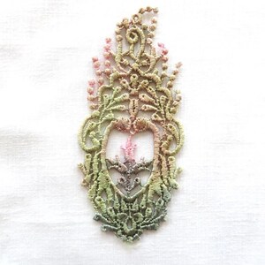 May include: A delicate, intricate lace design in shades of green, pink, and white. The lace is in the shape of a stylized flower or a floral motif.
