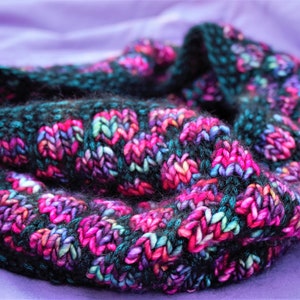 May include: A black, teal, pink and purple knitted infinity scarf. The scarf is made with a chunky yarn and has a textured stitch pattern.