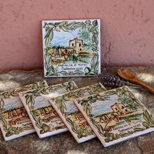 May include: A set of square ceramic coasters with a painted landscape scene, including a building, trees, and a blue sky. The coasters are decorated with olive branches and the text "FAMIGLIA E TUTTO Settembre 2025".