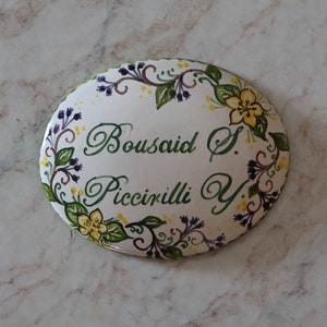 Op de afbeelding: Witte ovale keramische tegel met een groene, gele en paarse bloemenrand. De tegel heeft de namen "Bousaid S." en "Piccirilli Y." in cursief geschreven.