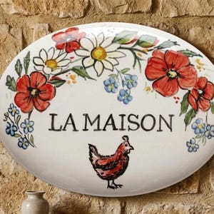Puede incluir: Letrero ovalado de cerámica con un diseño floral que presenta amapolas rojas, margaritas blancas y nomeolvides azules. El letrero dice "LA MAISON" e incluye una ilustración de pollo pintada a mano.