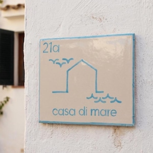 Pode incluir: Placa de número de casa quadrada em cerâmica com borda azul claro. A placa apresenta o número "21a" em azul claro, juntamente com uma ilustração de casa e ondas, e as palavras "casa di mare" em azul claro.