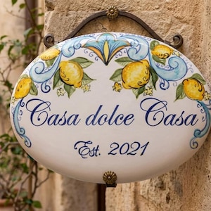 May include: Oval ceramic sign with a hand-painted design of lemons and blue swirls, with the words "Casa dolce Casa" and "Est. 2021" in navy blue script. The sign is mounted on a wrought iron bracket.