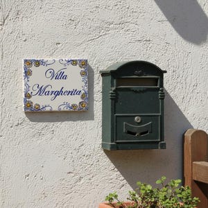 Puede incluir: Un buzón de metal verde oscuro y un letrero decorativo de cerámica con la inscripción "Villa Margherita" están montados en una pared blanca texturizada. El letrero tiene un borde floral en amarillo y azul.