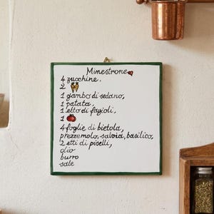 Può includere: Una targa da parete quadrata in ceramica bianca con bordo verde scuro, che riporta una ricetta italiana del Minestrone dipinta a mano in corsivo nero. Il testo include piccole illustrazioni di due carote, un pomodoro e un cuore rosso.