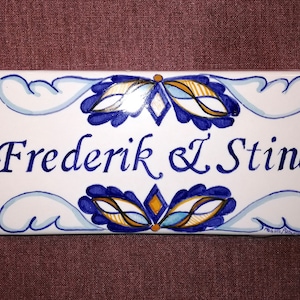 Può includere: Piastrella di ceramica bianca con disegni floreali blu e dorati. La piastrella ha il testo "Frederik & L Stine" scritto in corsivo blu.