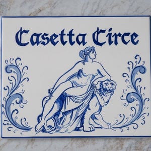 Puede incluir: Azulejo de cerámica blanco con ilustraciones y texto en azul. Presenta una ilustración clásica de una mujer reclinada sobre un león, con las palabras "Casette Circe" en una fuente decorativa. Diseños florales azules enmarcan la imagen.