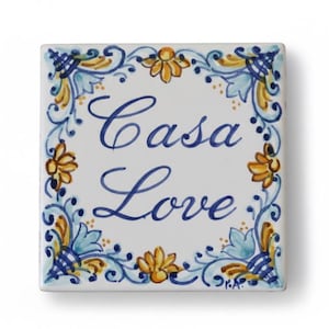 以下が含まれることがあります： 青い筆記体で「Casa Love」と書かれた正方形のセラミックタイル。タイルは白で、青、黄、金のフローラルとスクロールワークのデザインの装飾的なボーダーが付いています。装飾品です。