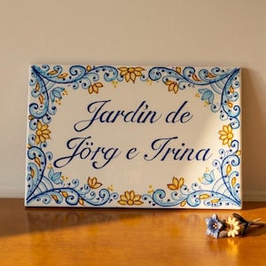 Op de afbeelding: Rechthoekige keramische plaat met de tekst "Jardin de Jörg e Trina" in donkerblauw schrift. De plaat is omrand met een bloemenontwerp in blauw, geel en wit. Een klein boeket bloemen ligt op een houten oppervlak.