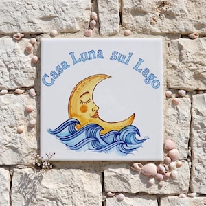 以下が含まれることがあります： ライトブルーの「Casa Luna sul Lago」の文字が入った正方形のセラミックタイル。青い波の上に、顔のある黄色の三日月が描かれています。タイルは貝殻に囲まれ、石壁に設置されています。
