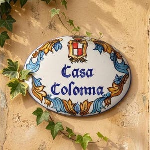 以下が含まれることがあります： 青い筆記体で「Casa Colonna」と書かれた楕円形のセラミックサイン。青と金のフローラル要素と上部の紋章が付いた装飾的なボーダーがあります。緑のツタがサインを囲んでいます。