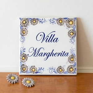 Könnte beinhalten: Quadratische weiße Keramikfliese mit den Worten "Villa Margherita" in eleganter blauer Schrift. Die Fliese ist mit einem Blumendesign mit gelben und weißen Gänseblümchen und blauen Akzenten umrandet. Zwei dekorative Knöpfe in Form von Gänseblümchen befinden sich im Vordergrund.