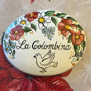 Op de afbeelding: Ovaal keramisch decoratief stuk met een witte basis, met de tekst "La Colombina" in zwart schrift, een duifillustratie en bloemmotieven in rood, wit en blauw. Het stuk is omgeven door een bloemenrand.