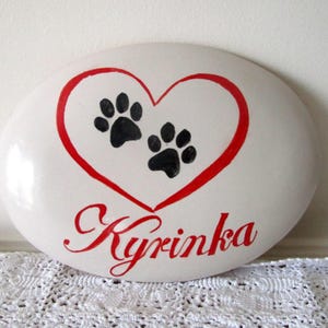 May include: Oval white stone with a red heart containing two black paw prints. The name "Kyrinka" is written in red script below. A decorative item, possibly a pet memorial or keepsake.