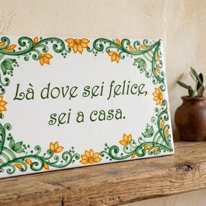 以下が含まれることがあります： イタリア語のフレーズ「Là dove sei felice, sei a casa」が書かれた長方形のセラミックタイル。タイルは白い背景に緑と黄色の花のボーダーがあります。近くには、緑の植物が入った小さな花瓶があります。